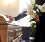 In schwarz gekleidete Frau trauert am Sarg mit Lilien in der Hand.