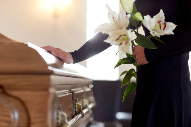 In schwarz gekleidete Frau trauert am Sarg mit Lilien in der Hand.